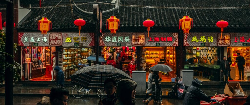 Fotografia em cores de lojas em mercado de Xangai. O dia está nublado, pessoas caminham sob guarda-chuvas. As lojas se destacam pela cores fortes de seus luminosos e lanternas típicas.