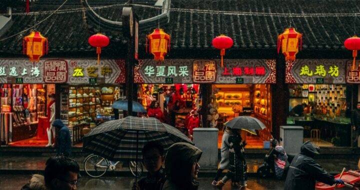 Fotografia em cores de lojas em mercado de Xangai. O dia está nublado, pessoas caminham sob guarda-chuvas. As lojas se destacam pela cores fortes de seus luminosos e lanternas típicas.