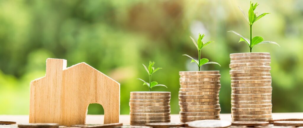 Fotografía en color. Modelo de casa de MDF y montones de monedas dispuestas en orden ascendente.