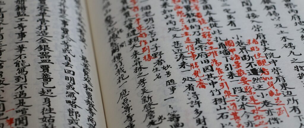 Fotografia colorida de livro escrito em chinês, com bonita caligrafia. Os caracteres são em tinta preta, com alguns em tinta vermelha ao lado.