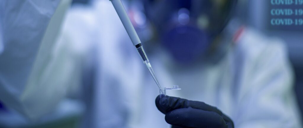 Foto em cores. Em primeiro plano, mãos enluvadas usam pipeta para colocar líquido dentro de pino. Ao fundo, o manipulador vestido em trajes de seguranças. Mais ao fundo, pode-se ler COVID-19 escrito 3 vezes.
