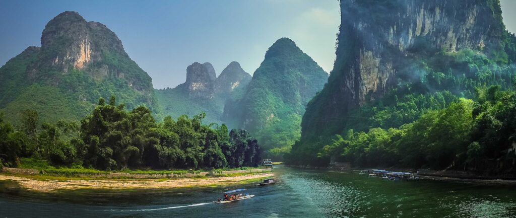 Fotografía panorámica en color. Día soleado, río con algunos botes, vegetación verde en las orillas, montañas altas al fondo.