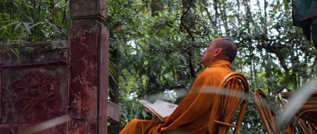 Fotografia em cores de monge budista sentado em uma cadeira, livro à mão, contemplando a natureza. Em Leschan, China.