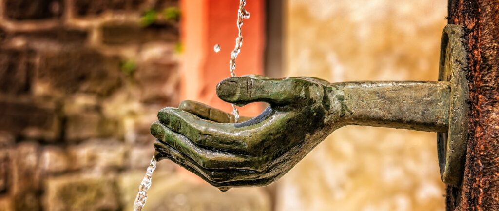 Fotografía en color de manos esculpidas formando un caparazón. Las manos parecen salir de una pared. Un hilo de agua cae sobre las manos y corre por debajo del