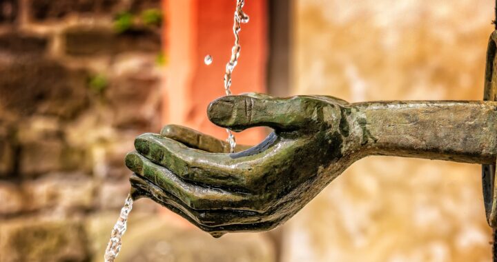Fotografía en color de manos esculpidas formando un caparazón. Las manos parecen salir de una pared. Un hilo de agua cae sobre las manos y corre por debajo del