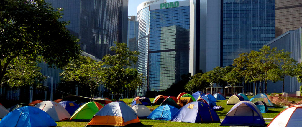Fotografia em cores de barracas em gramado ao lado do Conselho Legislativo de Hong Kong. Imagem representa os protestos Occupy Central, de 2014.