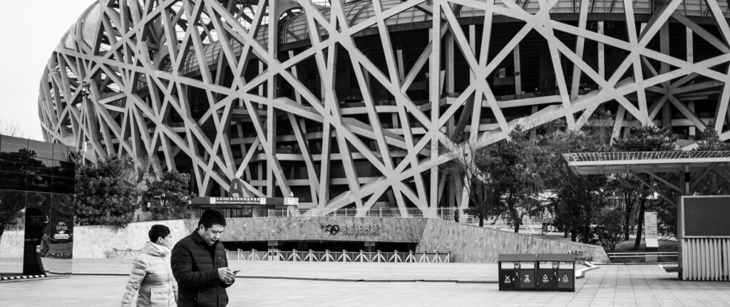 Fotografía en blanco y negro de una pareja caminando frente al Estadio Nido de Pájaro en Beijing. En primer plano, el hombre mira un celular.