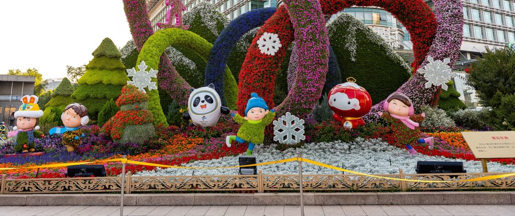 Fotografia em cores de decoração celebrando as Olimpíadas de Inverno em Pequim 2022. Além de flores, a decoração traz estátuas dos mascotes dos jogos e de crianças estilizadas.