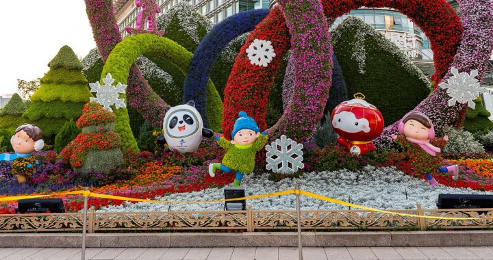 Fotografia em cores de decoração celebrando as Olimpíadas de Inverno em Pequim 2022. Além de flores, a decoração traz estátuas dos mascotes dos jogos e de crianças estilizadas.