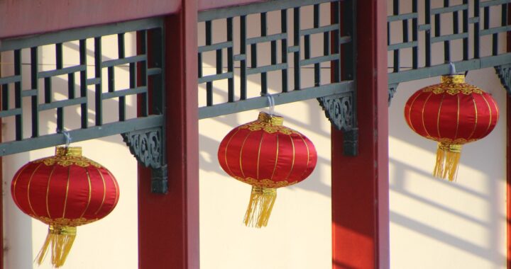 Fotografía en color de tres faroles chinos rojos y dorados. Cuelgan de un pórtico de madera pintado de rojo y decorado con recortes en material negro.
