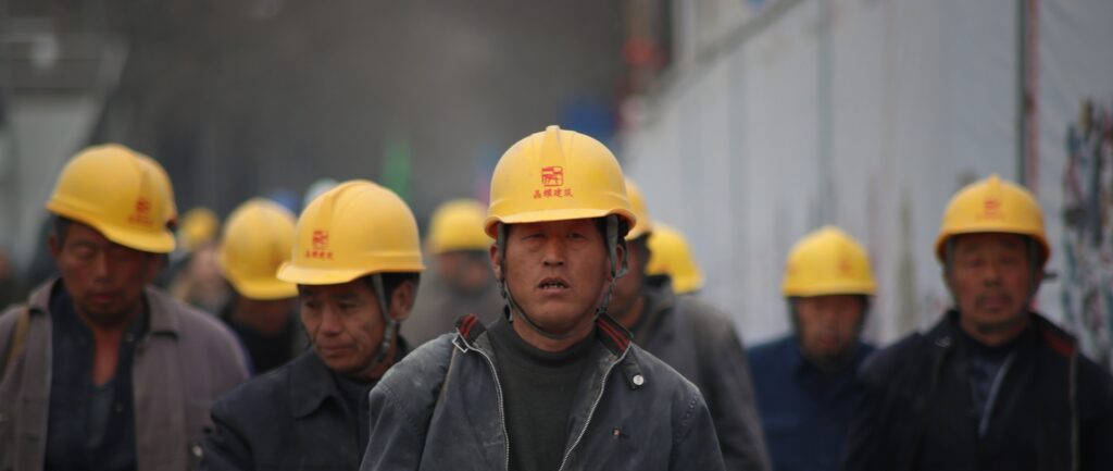 Fotografía en color de trabajadores industriales chinos. Visten trajes grises y cascos amarillos.