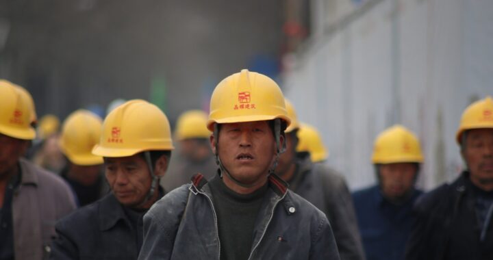Fotografía en color de trabajadores industriales chinos. Visten trajes grises y cascos amarillos.