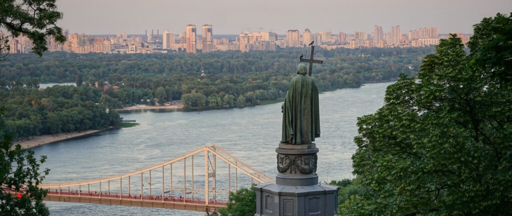 Fotografía panorámica en color de Kiev, Ucrania. En primer plano, la estatua de Vladimir el Grande, vista desde atrás. Al fondo, un puente atirantado sobre un río, muchos árboles y, más lejos, edificios.