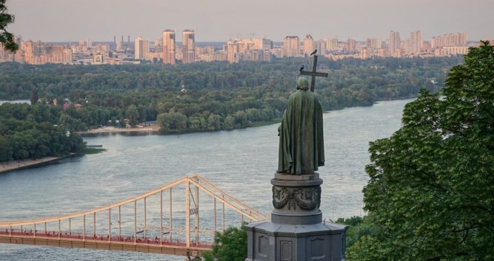 Fotografía panorámica en color de Kiev, Ucrania. En primer plano, la estatua de Vladimir el Grande, vista desde atrás. Al fondo, un puente atirantado sobre un río, muchos árboles y, más lejos, edificios.
