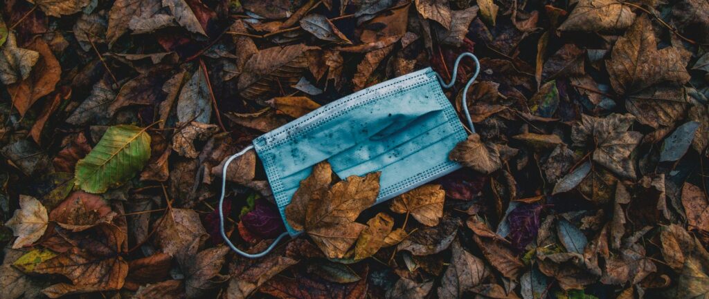 Fotografía en color de una mascarilla quirúrgica azul abandonada en un suelo cubierto de hojas secas.