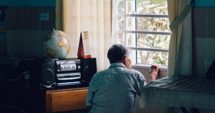 Fotografia em cores de homem idoso, aparentemente chinês. Ele está de costas, sentado dentro de casa, olhando para fora, pela janela.