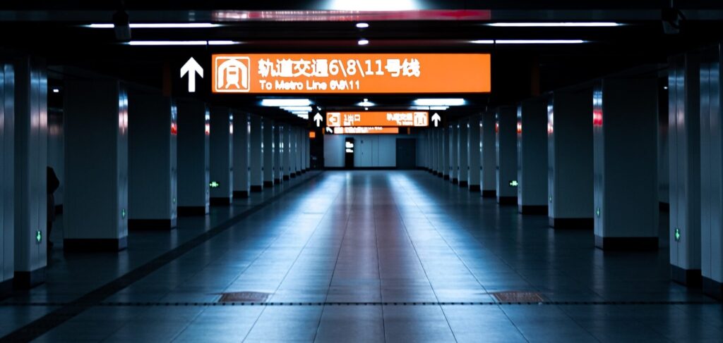 Fotografia em cores de estação de metrô de Shanghai. O corredor parece vazio, há apenas uma placa de direção iluminada, nas cores laranja e branca.