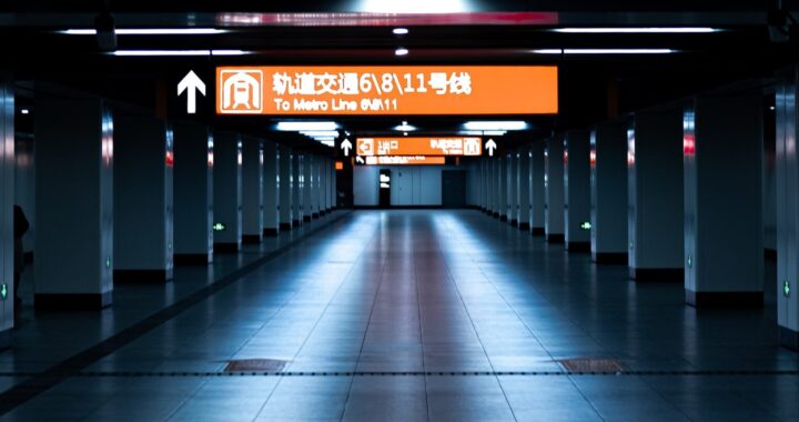 Fotografia em cores de estação de metrô de Shanghai. O corredor parece vazio, há apenas uma placa de direção iluminada, nas cores laranja e branca.
