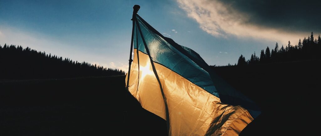 Fotografía en color de la bandera ucraniana contra la luz del sol en un ambiente sombrío al aire libre. Al fondo, árboles y nubes.