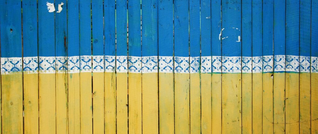 Fotografia em cores de portão feito de ripas de madeira e pintado com as cores azul e amarela, da bandeira da Ucrânia. Entre as duas cores, um padrão decorativo em branco.