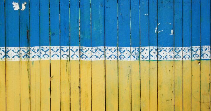 Fotografia em cores de portão feito de ripas de madeira e pintado com as cores azul e amarela, da bandeira da Ucrânia. Entre as duas cores, um padrão decorativo em branco.