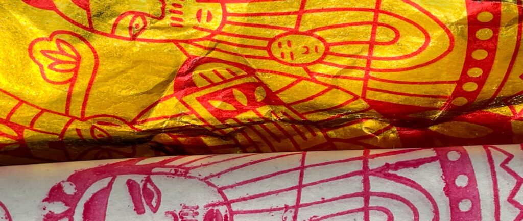 Fotografía en color del papel de incienso utilizado como ofrenda al difunto. Uno de ellos es blanco, el otro es amarillo, ambos tienen diseños rojos. Los dibujos son de entidades masculinas.