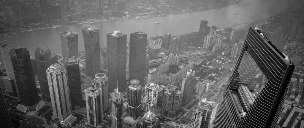 Fotografia em branco e preto de vista aérea de Xangai, onde se vê prédios do distrito de Huangpu em primeiro plano, com destaque para o Shanghai World Financial Center. Ao fundo, vê-se o rio e um pedaço do Bund.