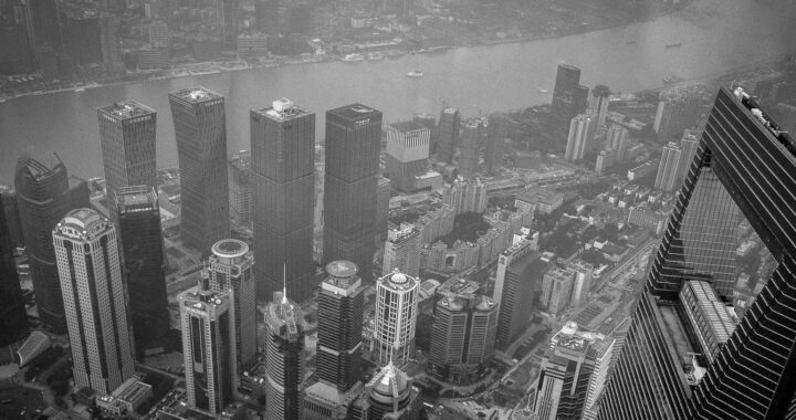 Fotografia em branco e preto de vista aérea de Xangai, onde se vê prédios do distrito de Huangpu em primeiro plano, com destaque para o Shanghai World Financial Center. Ao fundo, vê-se o rio e um pedaço do Bund.