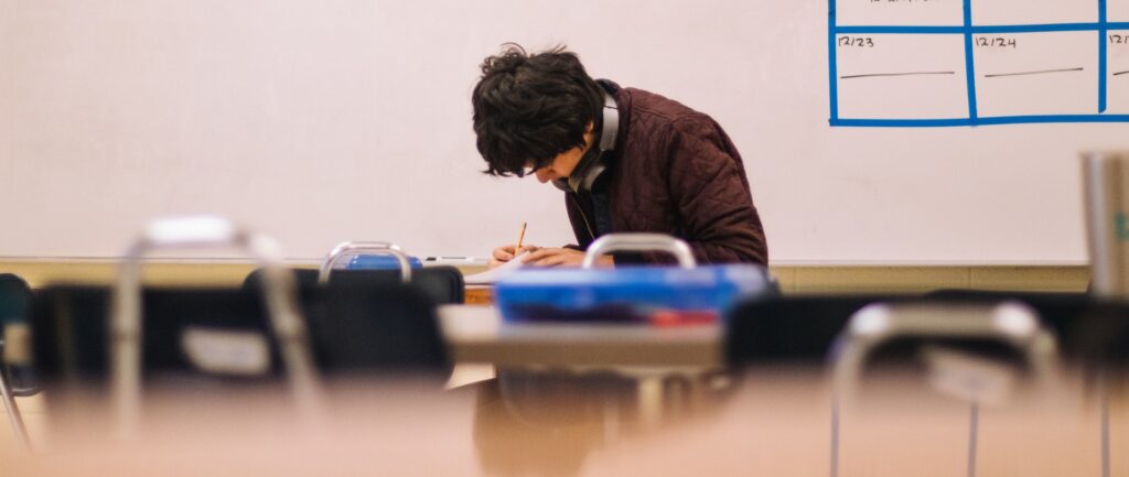 Fotografía en color de un adulto joven escribiendo en un cuaderno dentro del aula.
