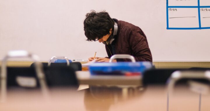 Fotografía en color de un adulto joven escribiendo en un cuaderno dentro del aula.