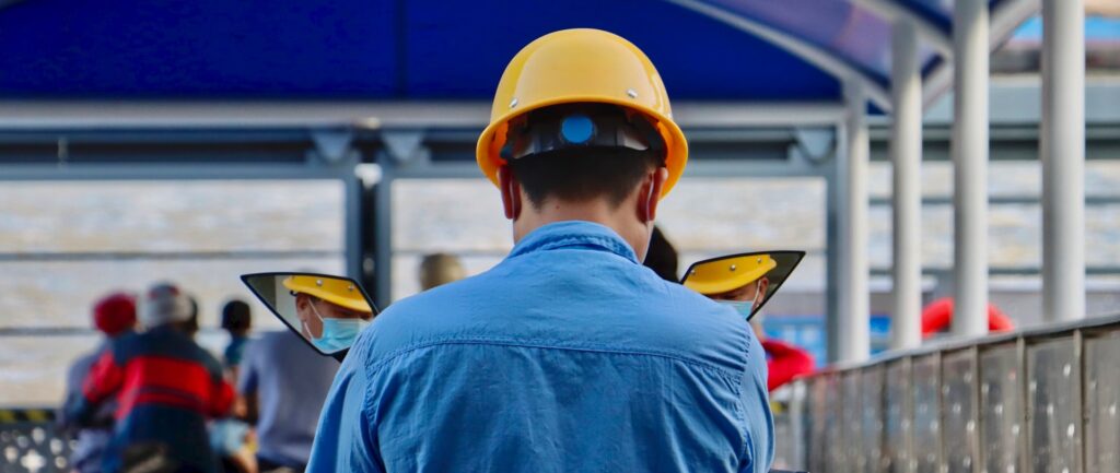 Fotografia em cores de trabalhador chinês usando capacete amarelo. Ele está de costas, vestindo uma camisa azul, e é possível ver parte de seu rosto (com máscara facial) pelo reflexo em dois retrovisores de sua moto. Ao fundo, outros trabalhadores, sentados em motocicletas.