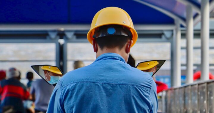 Fotografia em cores de trabalhador chinês usando capacete amarelo. Ele está de costas, vestindo uma camisa azul, e é possível ver parte de seu rosto (com máscara facial) pelo reflexo em dois retrovisores de sua moto. Ao fundo, outros trabalhadores, sentados em motocicletas.
