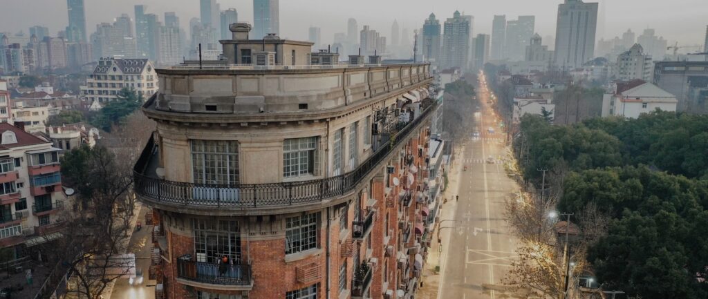 Fotografia em cores de um prédio de esquina em uma intersecção de Xangai. Vê-se pouco movimento nas ruas a seu redor e diversos prédios ao fundo.