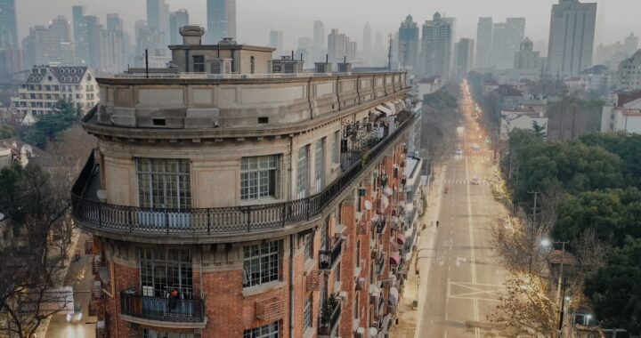 Fotografia em cores de um prédio de esquina em uma intersecção de Xangai. Vê-se pouco movimento nas ruas a seu redor e diversos prédios ao fundo.