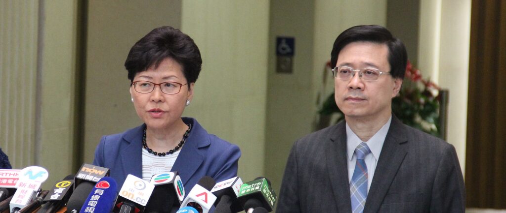 Fotografia em cores de Carrie Lam e John Lee dando entrevista. À frente deles, diversos microfones.