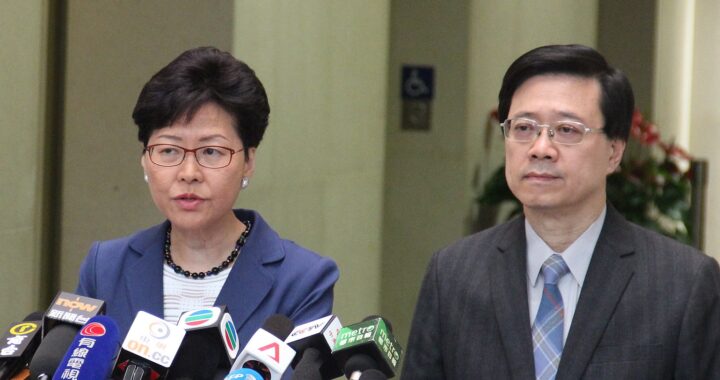 Fotografia em cores de Carrie Lam e John Lee dando entrevista. À frente deles, diversos microfones.