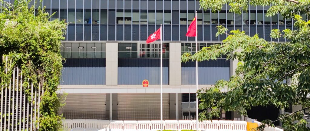 Fotografía en color de la fachada del edificio del Consejo Legislativo de Hong Kong. En el frente se puede ver una bandera china y una bandera de Hong Kong. La bandera de hong kong está en un poste inferior. En el edificio se puede ver el escudo de armas chino.