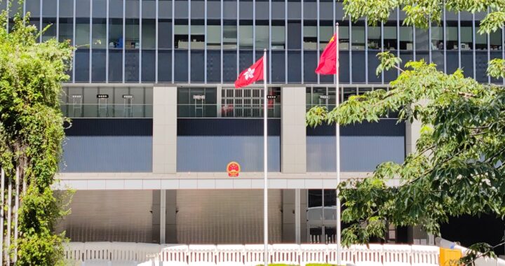 Fotografía en color de la fachada del edificio del Consejo Legislativo de Hong Kong. En el frente se puede ver una bandera china y una bandera de Hong Kong. La bandera de hong kong está en un poste inferior. En el edificio se puede ver el escudo de armas chino.