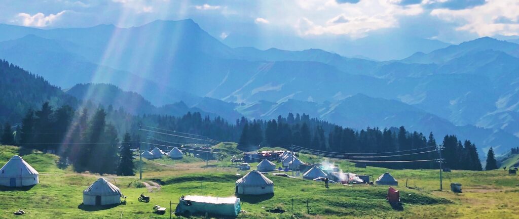 Fotografia em cores de cenário idílico em Xinjiang: céu azul, raios de sol, cabanas rústicas sobre gramado verde. Também é possível ver postes e cabos de energia, além de uma pessoa.