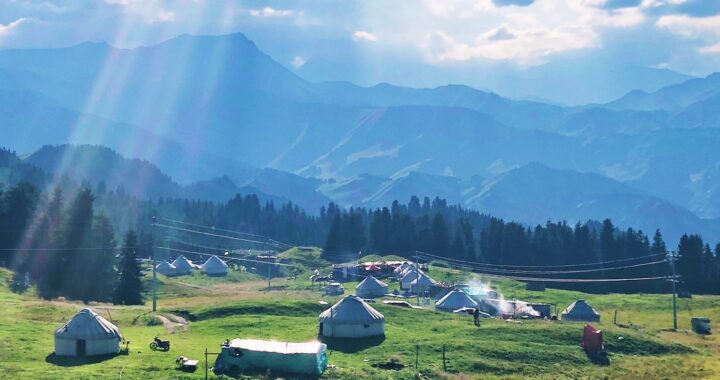 Fotografia em cores de cenário idílico em Xinjiang: céu azul, raios de sol, cabanas rústicas sobre gramado verde. Também é possível ver postes e cabos de energia, além de uma pessoa.