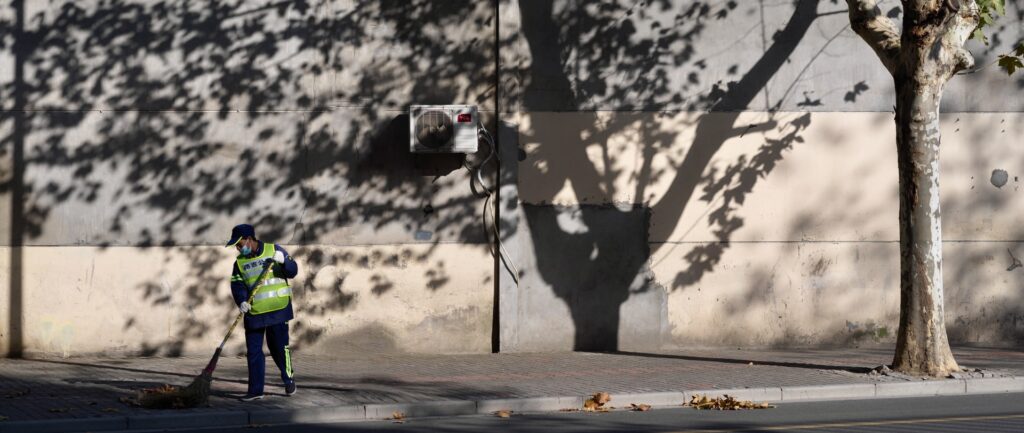 Fotografia em cores de rua vazia em Shanghai. Há apenas um faxineiro de máscara valendo o chão, com uma árvore ao seu lado. Ao fundo, vê-se a sombra da árvore em uma parede.