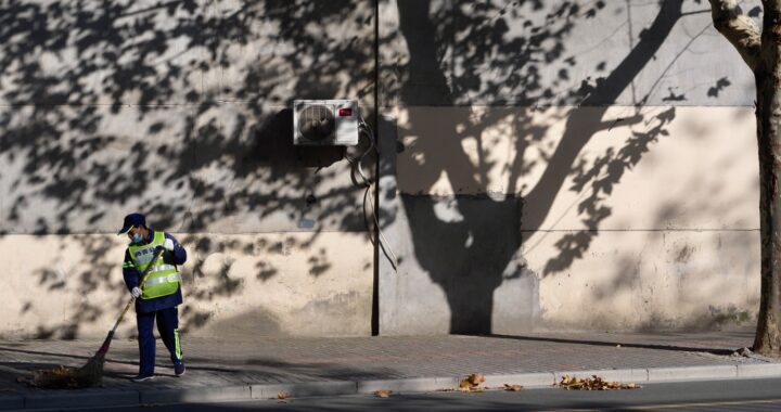 Fotografia em cores de rua vazia em Shanghai. Há apenas um faxineiro de máscara valendo o chão, com uma árvore ao seu lado. Ao fundo, vê-se a sombra da árvore em uma parede.