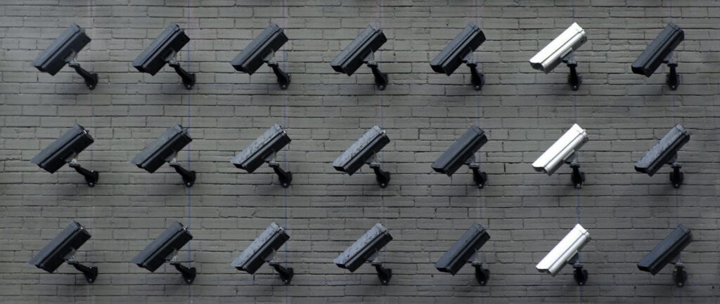 Fotografia de diversas câmeras de segurança, algumas pretas, outras brancas, em uma parede. Todas estão viradas para a mesma direção, esquerda abaixo.