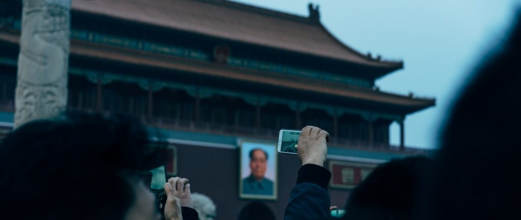 Fotografia em cores da Porta da Paz Celestial. Em primeiro plano, uma mão ergue um celular. Ao fundo vê-se o retrato de Mao.