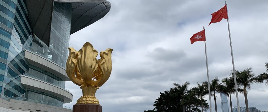 Fotografia em cores do Golden Bauhinia Square, onde ocorrem celebrações da transferência de soberania de Hong Kong. Vê-se a flor-símbolo de Hong Kong, uma escultura dourada, à frente; ao fundo, as bandeiras da China e de Hong Kong.