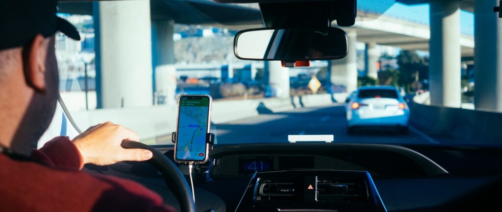 Fotografia em cores de motorista dirigindo entre viadutos guiado por um aplicativo no celular.