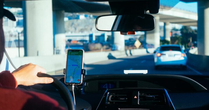 Fotografia em cores de motorista dirigindo entre viadutos guiado por um aplicativo no celular.