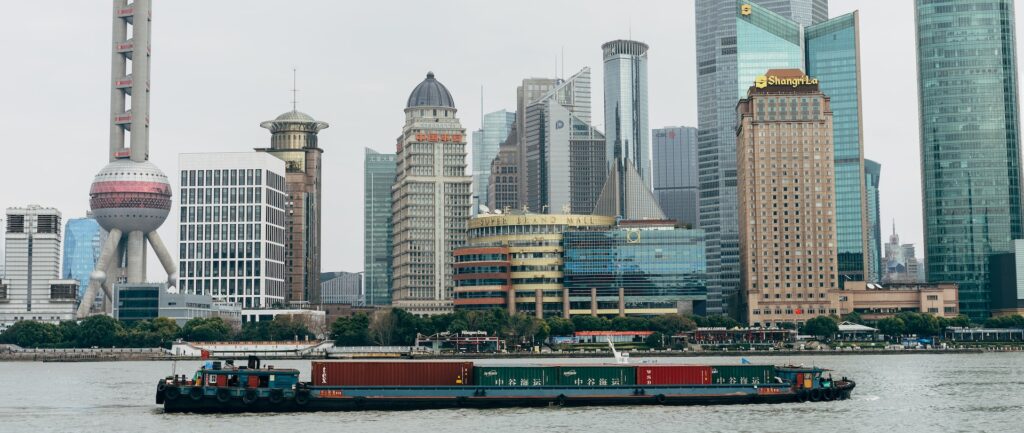 Fotografia em cores de barco com conteiners passando pelo rio Huangpu, com prédios icônicos de Shanghai ao fundo.