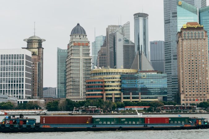 Fotografia em cores de barco com conteiners passando pelo rio Huangpu, com prédios icônicos de Shanghai ao fundo.