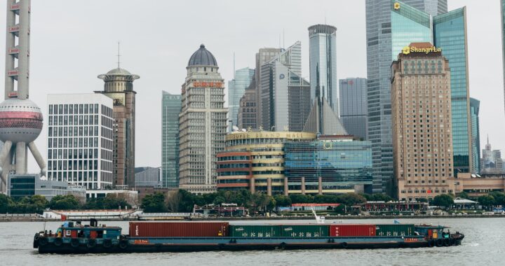 Fotografia em cores de barco com conteiners passando pelo rio Huangpu, com prédios icônicos de Shanghai ao fundo.